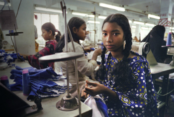 A girl works in a sweatshop, sewing garments. Large numbers of people are enslaved or at least unethically employed. Many products produced by such labor are cheap in price for the consumer, but expensive for the worker.